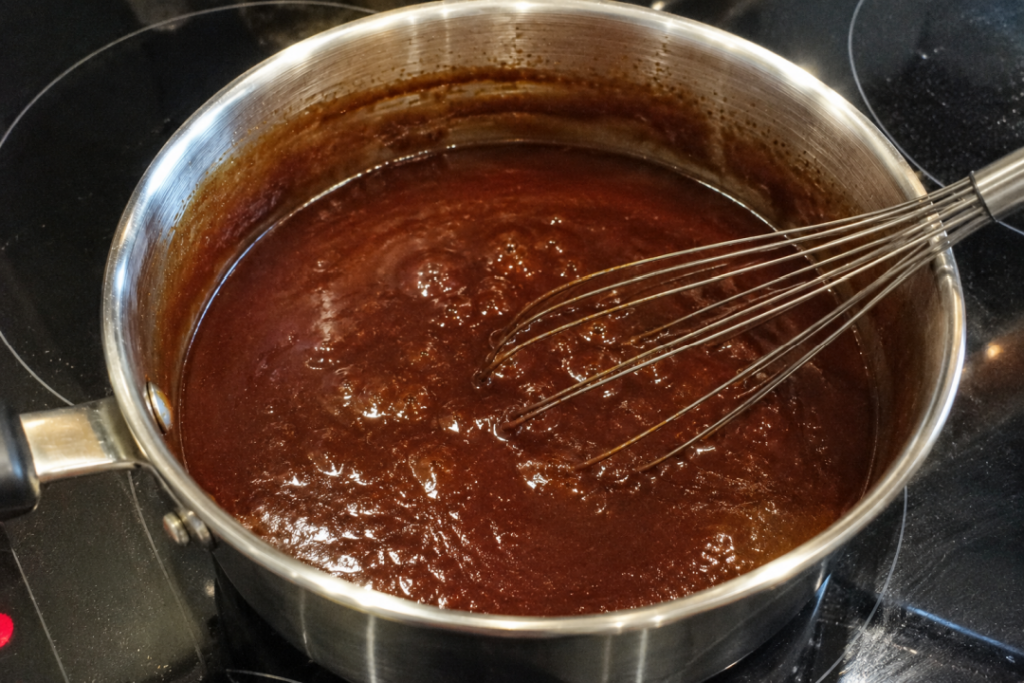 Homemade BBQ sauce gently simmering in a stainless steel saucepan on a black glass cooktop with a metal whisk resting in the thick, bubbling sauce.