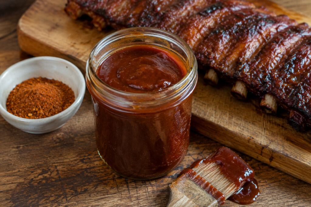 Homemade BBQ sauce in a glass mason jar with a sauce-covered basting brush, dry barbecue seasoning, and glazed pork ribs on a rustic wooden table.