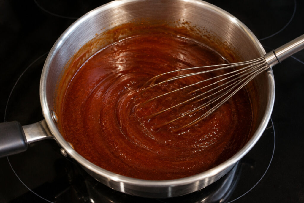 Homemade BBQ sauce being whisked in a stainless steel saucepan on a black glass cooktop until smooth and well combined.