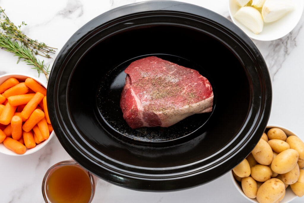 Rump roast seasoned with salt and pepper in a slow cooker with potatoes, carrots, onion, fresh rosemary, thyme, and beef broth ready for cooking.