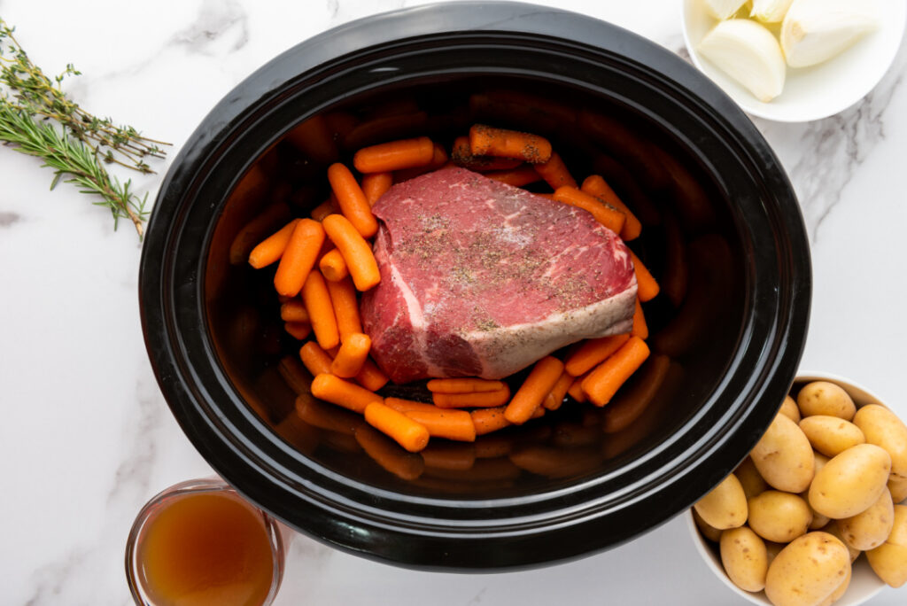 Baby carrots added around a seasoned rump roast in a slow cooker before cooking.