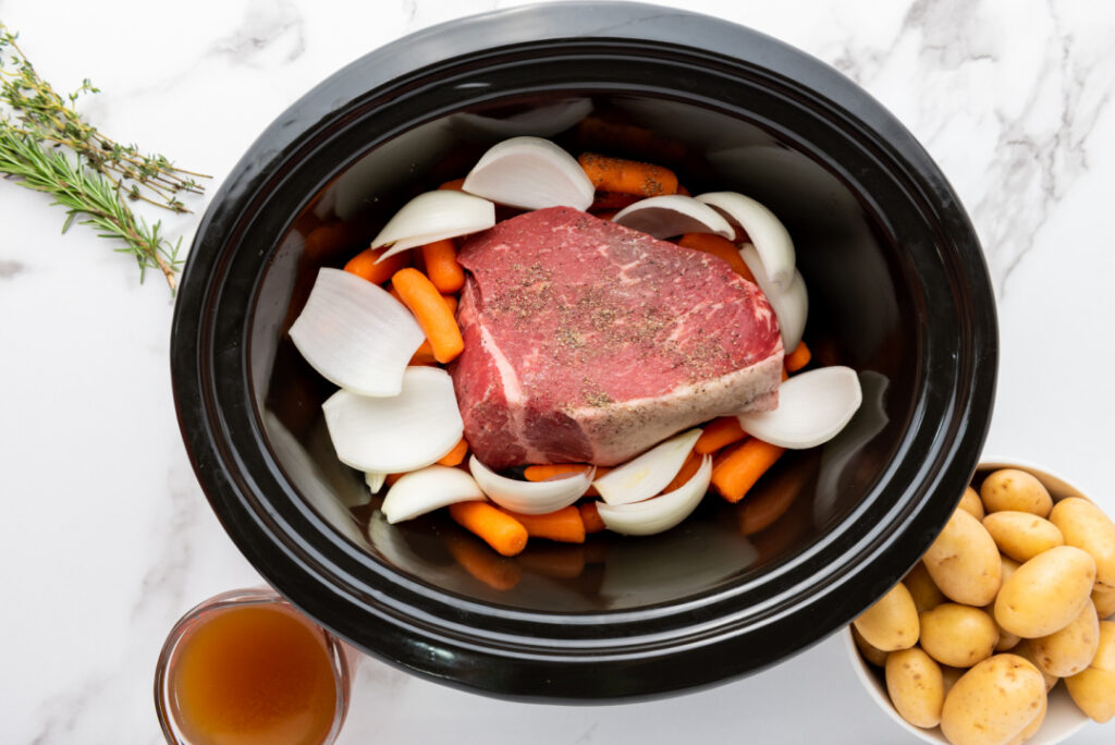 Quartered yellow onion added to a seasoned rump roast and carrots in a slow cooker before cooking.