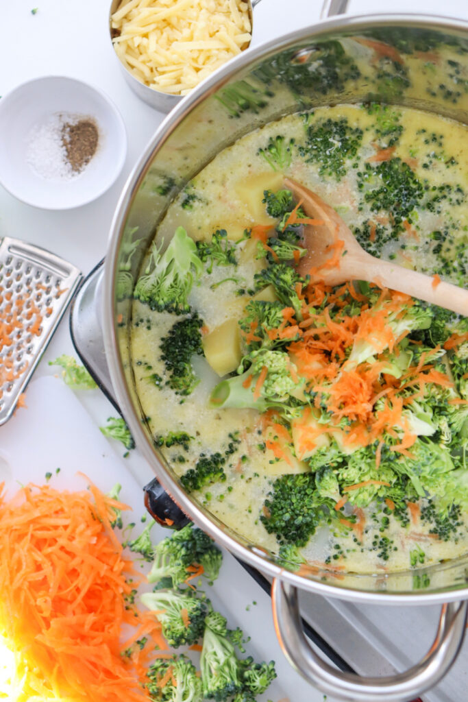 Fresh broccoli florets and grated carrots being added to homemade Broccoli Cheddar Soup before simmering until tender.