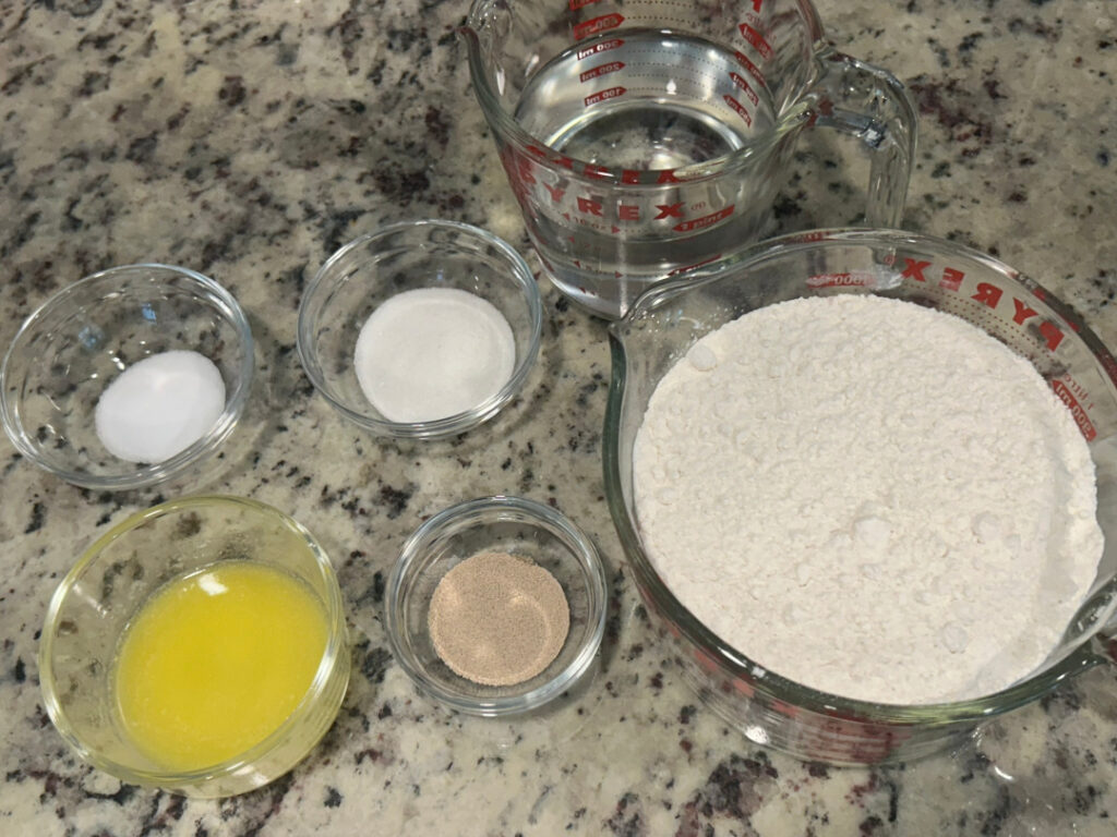 Measured ingredients for homemade basic white bread including all-purpose flour, warm water, melted butter, active dry yeast, sugar, and salt arranged on a kitchen countertop before mixing.