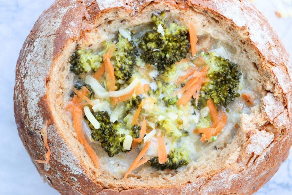 Close-up of creamy homemade Broccoli Cheddar Soup served in a crusty sourdough bread bowl with broccoli, carrots, potatoes, and freshly shredded sharp cheddar cheese.
