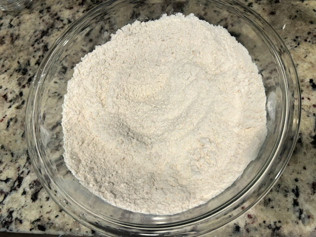 The dry ingredients for homemade basic white bread, including all-purpose flour, active dry yeast, sugar, and salt, mixed together in a large glass bowl before adding the warm water and melted butter.