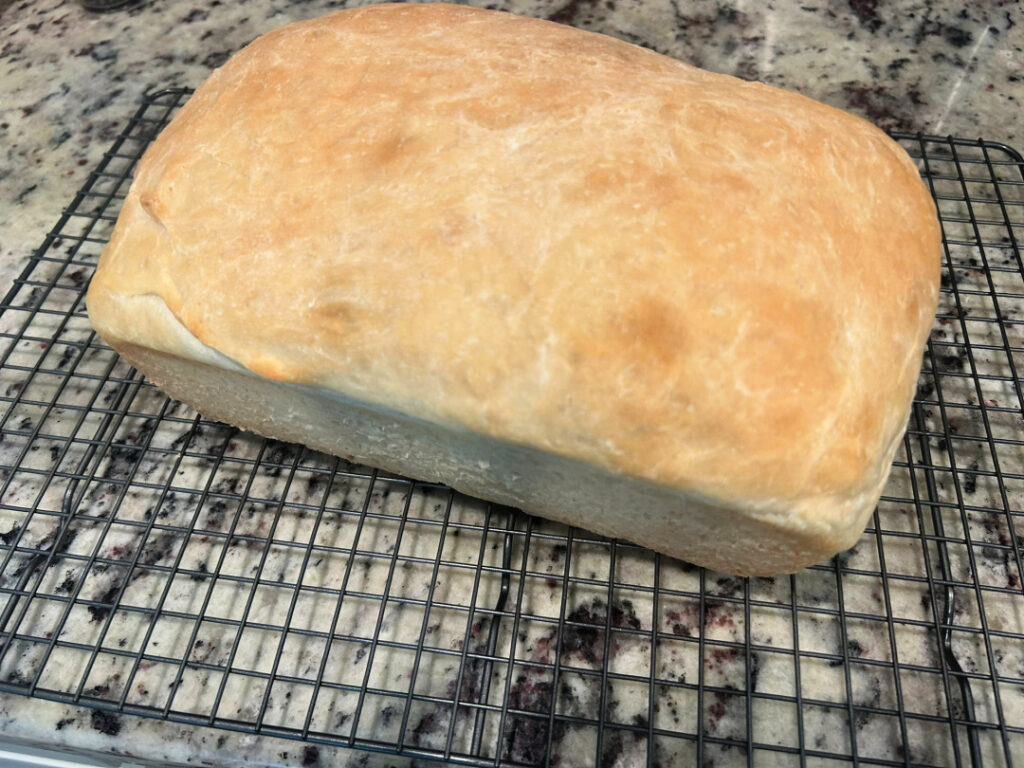 A freshly baked loaf of homemade basic white bread with a golden brown crust cooling on a wire rack after baking.
