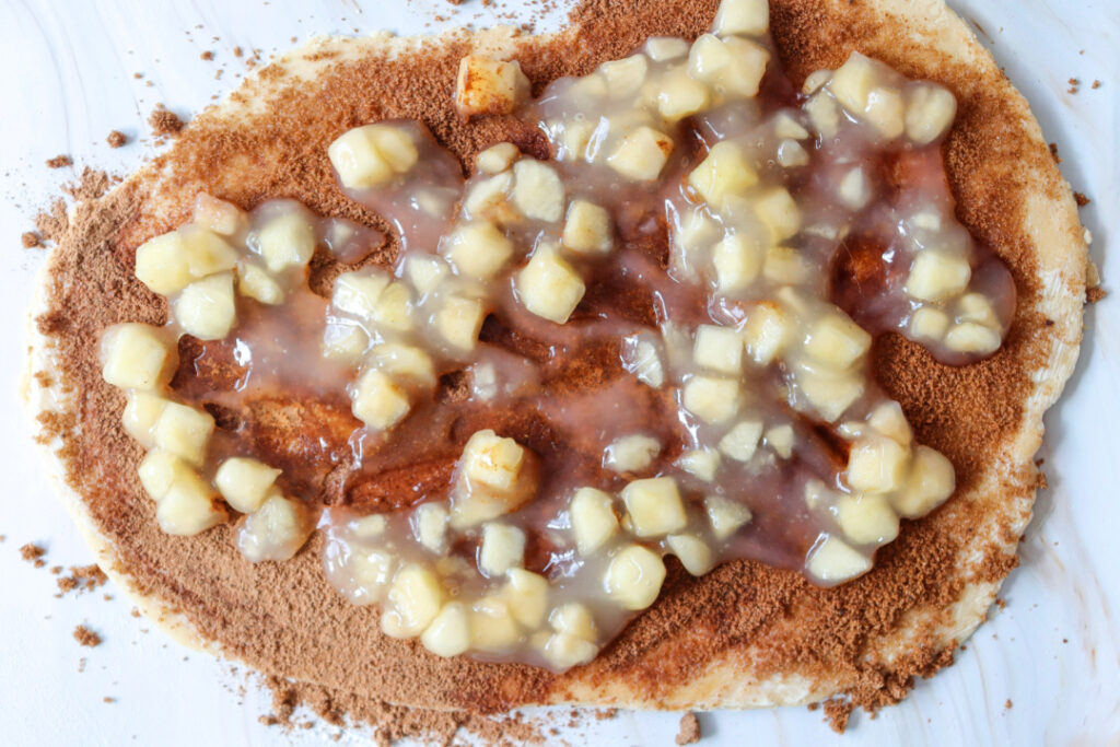 Store-bought apple pie filling spread over cinnamon and brown sugar on rolled dough for apple pie cinnamon rolls.