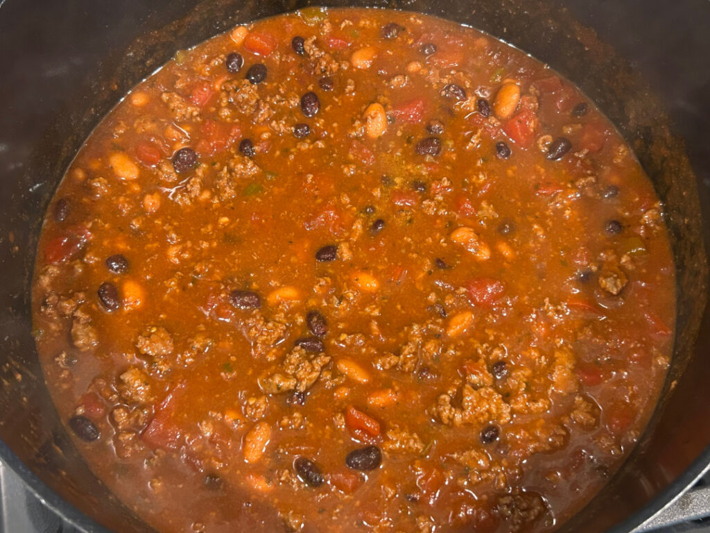 Beef and bean chili with ground beef, pinto beans, black beans, and diced tomatoes simmering in a large pot.