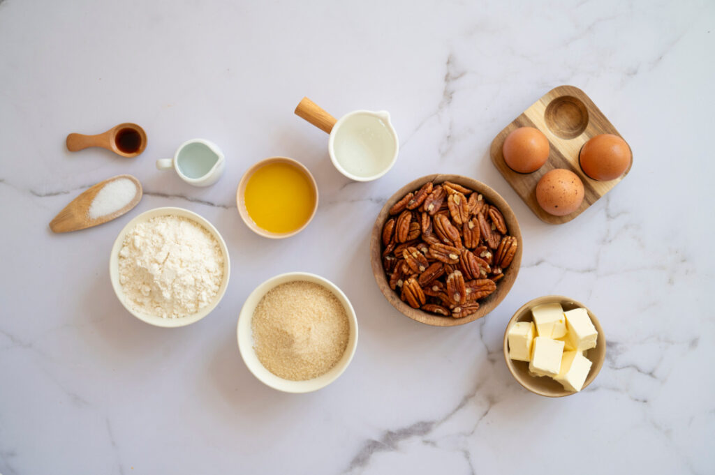 Ingredients for classic pecan pie arranged on a marble surface, including pecans, eggs, butter, brown sugar, flour, corn syrup, vanilla, and salt.