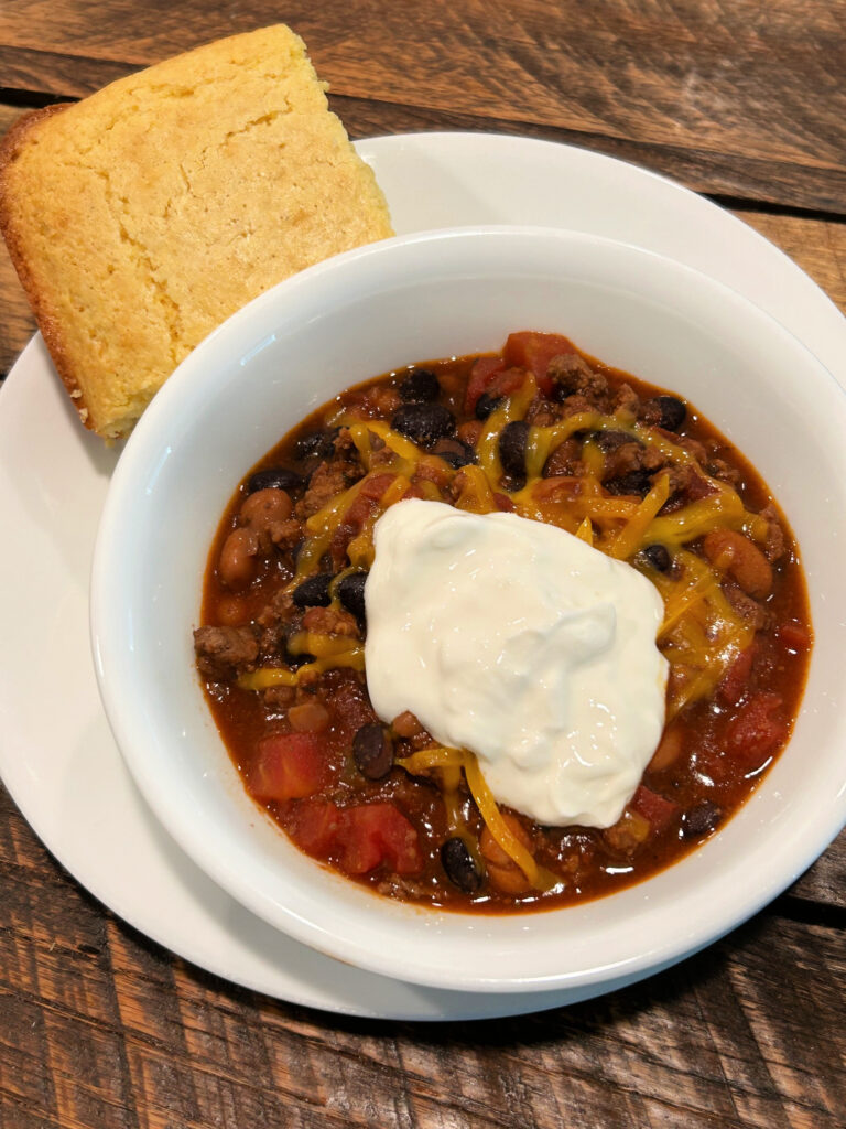 Bowl of easy beef and bean chili topped with shredded cheddar cheese and sour cream and served with cornbread.