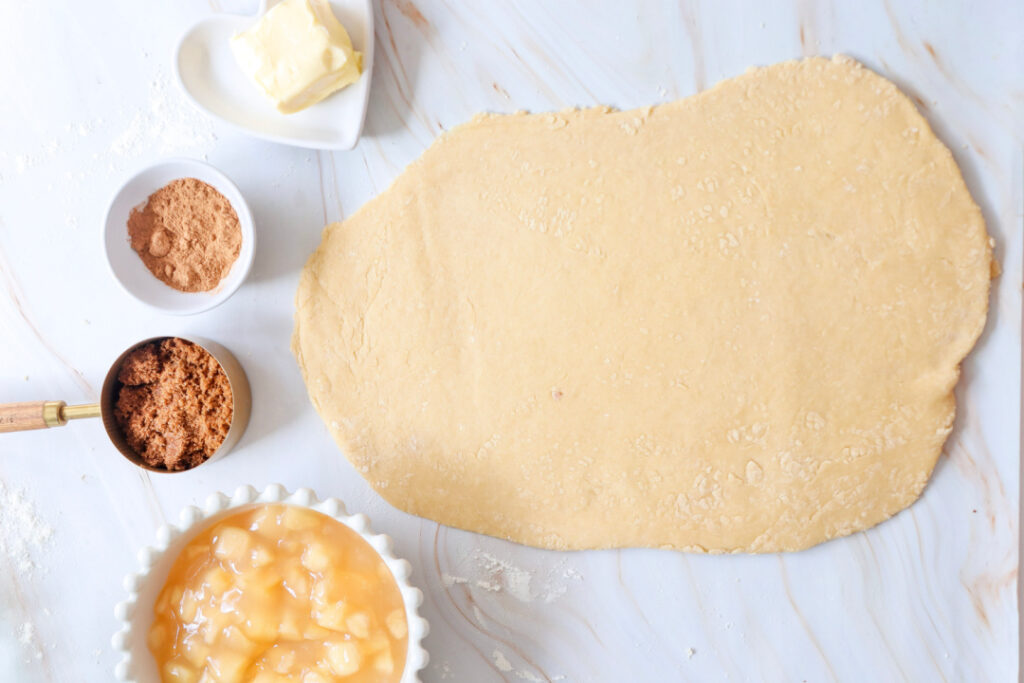 Homemade apple pie cinnamon roll dough rolled into a rectangle with apple pie filling, brown sugar, cinnamon, and softened butter ready to add.