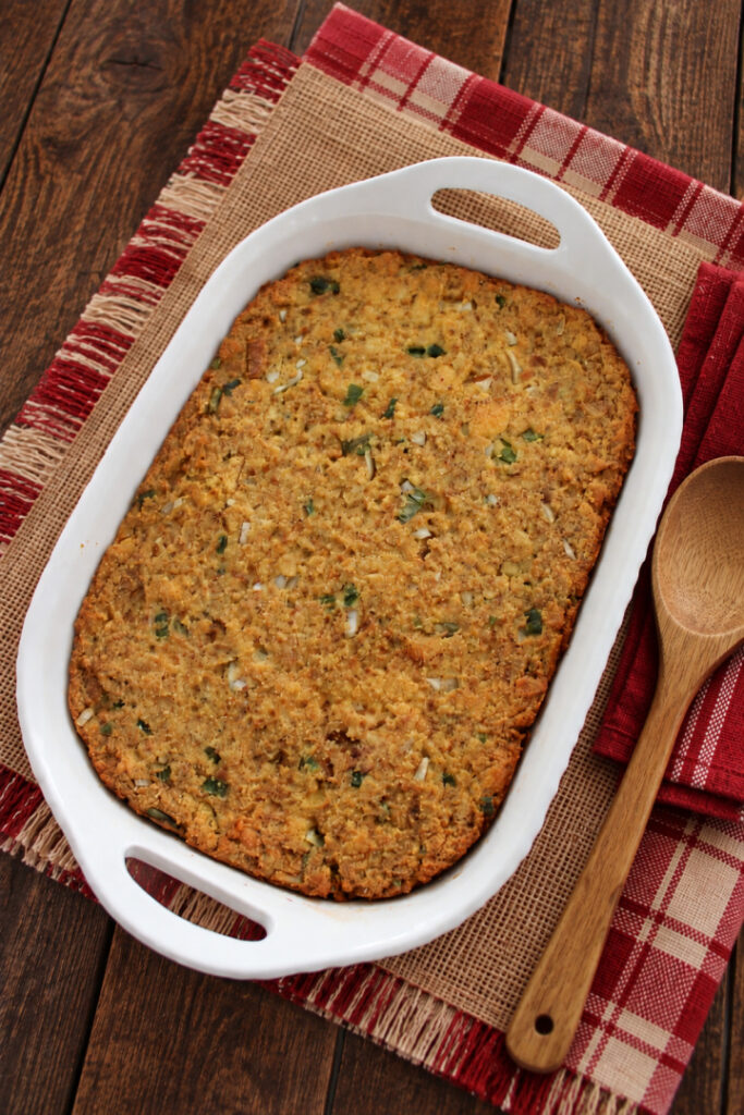 Southern cornbread dressing in a white casserole dish on a rustic wooden table with red plaid linens and a wooden serving spoon.